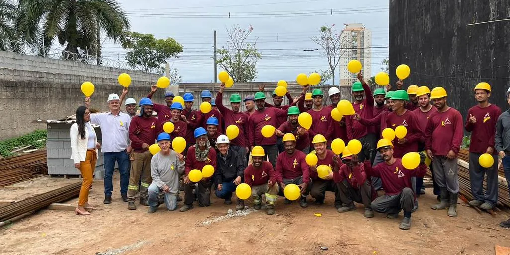 Equipe de obra com capacetes e balões amarelos reunida em campanha de saúde e segurança